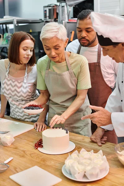 Mujer madura en delantal decorando pastel con bayas junto a sus amigos interracial y chef - foto de stock