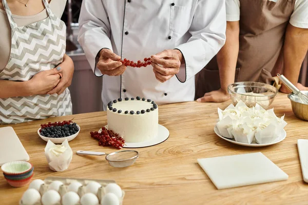 Обрізаний вид зрілого шеф-кухаря, який прикрашає смачний торт з червоною смородиною поруч зі своїми різноманітними студентами — стокове фото