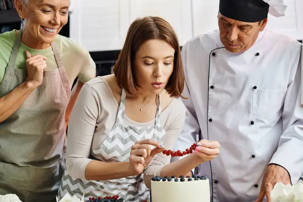 Молодая женщина в фартуке украшает торт красной смородиной рядом с ее веселым зрелым другом и шеф-поваром — стоковое фото