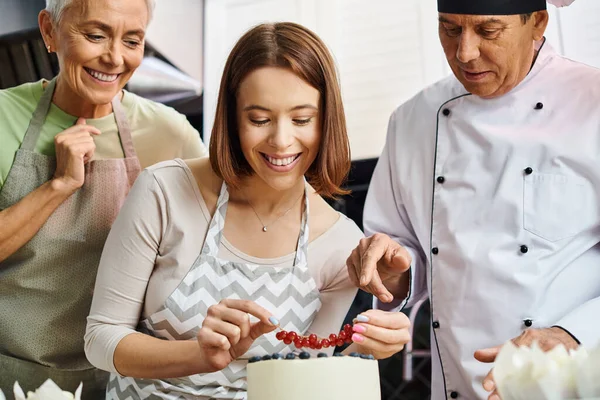 Joyeuse jeune femme décoration gâteau avec des baies à côté de son ami joyeux et chef mature — Photo de stock