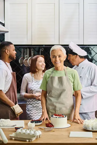 Focus sur femme mûre avec gâteau souriant à la caméra avec des amis divers flous et chef sur toile de fond — Photo de stock