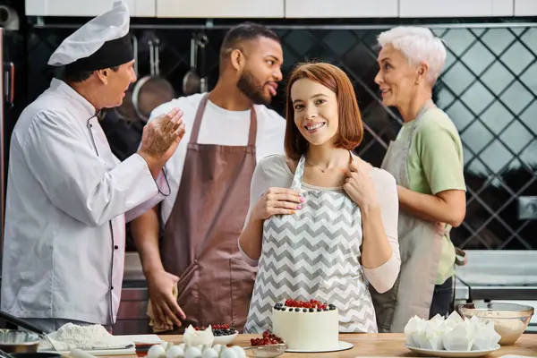Jeune femme joyeuse dans tablier regardant la caméra à côté du gâteau avec ses amis joyeux et divers sur fond — Photo de stock