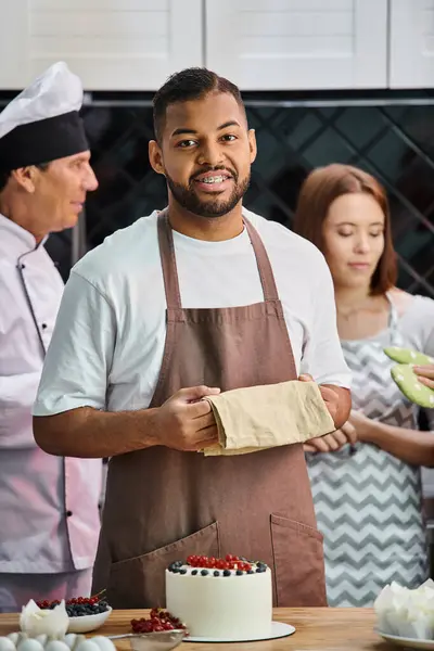 Enfoque en el hombre afroamericano feliz sonriendo a la cámara con su borroso amigo y chef en el telón de fondo - foto de stock