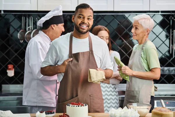Africano americano allegro uomo accanto a torta sorridente a macchina fotografica con chef e i suoi amici su sfondo — Foto stock