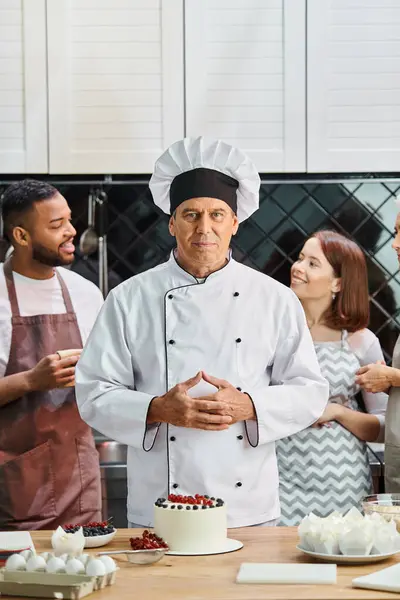 Красивый зрелый шеф-повар в белой шляпе с тортом на столе, смотрящий в камеру рядом со своими студентами — стоковое фото