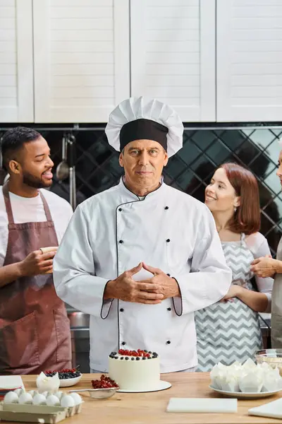 Handsome mature chef in white hat with cake on table looking at camera next to his students — Stock Photo