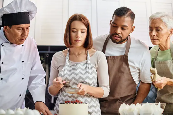 Gut aussehende multikulturelle Studenten und reifer Koch beobachten junge schöne Frau beim Dekorieren von Kuchen — Stockfoto