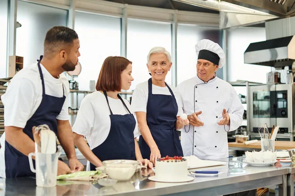 Handsome mature chief cook in white hat talking to his diverse jolly chefs in aprons, confectionary — Stock Photo