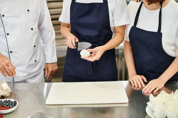 Обрезанный вид зрелой женщины, разбивающей яйцо ножом рядом со своими коллегами, кондитерская — стоковое фото