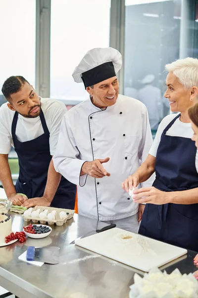 Alegre maduro jefe cocinero sonriendo a su interracial chefs mientras ellos hornear juntos, confitería - foto de stock