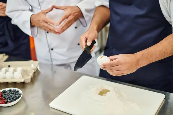 Vista recortada del chef afroamericano en delantal azul rompiendo huevo con cuchillo junto a sus colegas - foto de stock