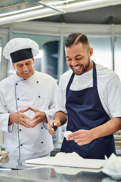 Jovem chef americano africano alegre em avental azul quebrando ovo ao lado de seu cozinheiro chefe alegre — Fotografia de Stock