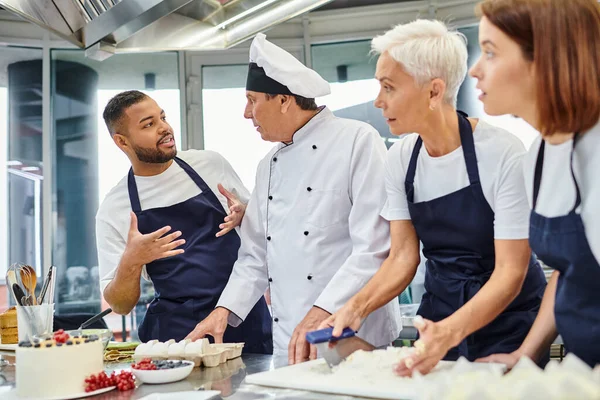 Gioioso chef afroamericano in grembiule parlando con i suoi colleghi mentre lavorano con la pasta — Foto stock