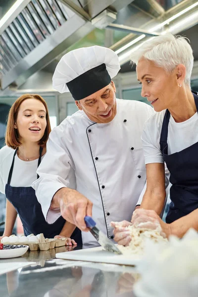 Joyeux chef cuisinier mature en chapeau blanc aidant ses jolies femmes chefs avec de la pâte, confiserie — Photo de stock