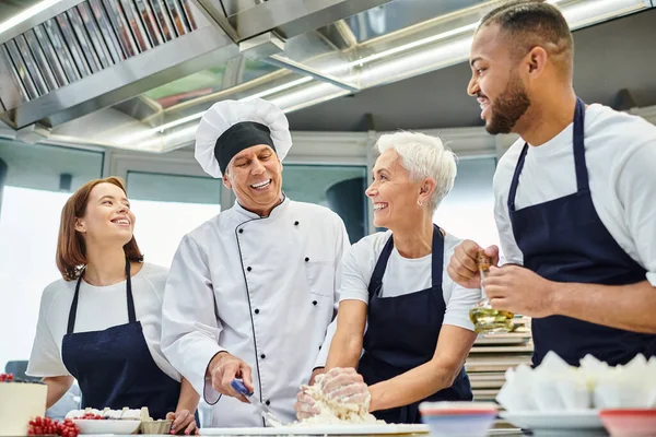 Alegre cocinero jefe maduro en sombrero blanco ayudando a sus chefs multiculturales con masa, confitería - foto de stock