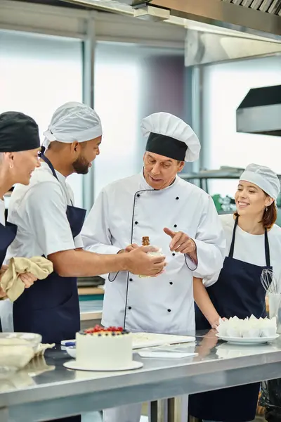 Mature good looking chief cook in white hat helping his interracial chefs with dough, confectionary — Stock Photo