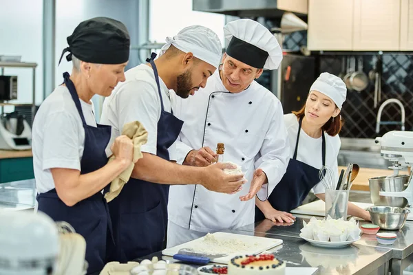 Bom olhar cozinheiro chefe maduro ajudando seus chefs multiculturais em toques com massa, confeitaria — Fotografia de Stock