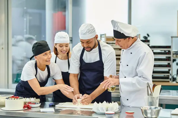 Chefs multiculturales alegres en delantales azules trabajando junto con la masa al lado del cocinero jefe - foto de stock