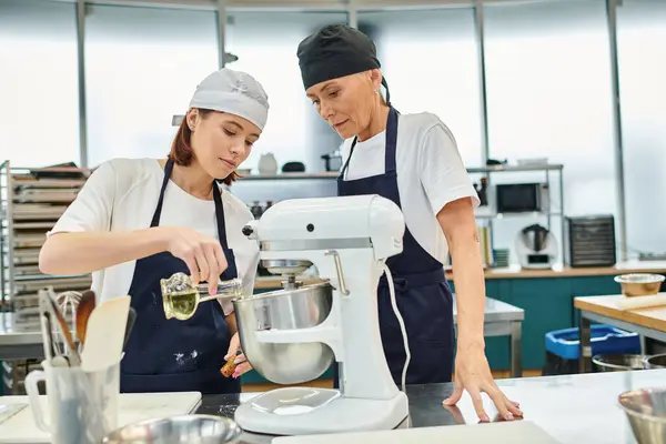 Attractive female chefs in blue aprons and toques adding a little bit of oil to mixer, confectionery — Stock Photo