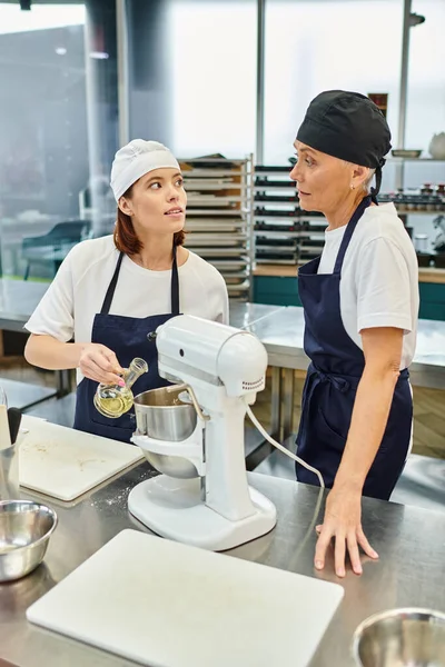 Красиві жіночі шеф-кухарі в синіх фартухах і тосках говорять, додаючи олію в міксер, кондитерські вироби — стокове фото