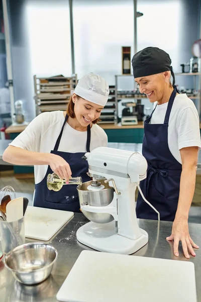 Chef femminili allegre in grembiuli e toques sorridenti felicemente e aggiungendo olio nel mixer, dolciaria — Foto stock