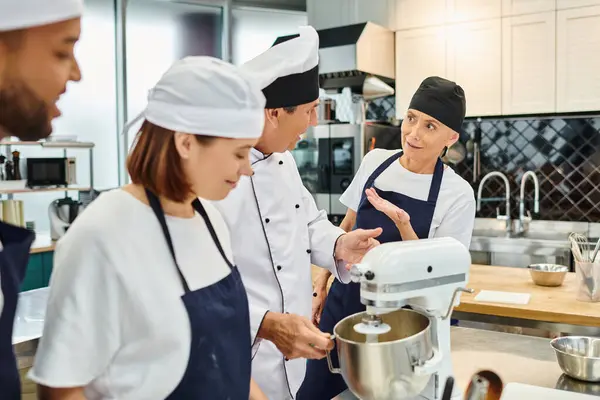 Chefs joyeux multiraciaux dans des tabliers bleus et des toques parler avec leur chef cuisinier à côté du mélangeur — Photo de stock