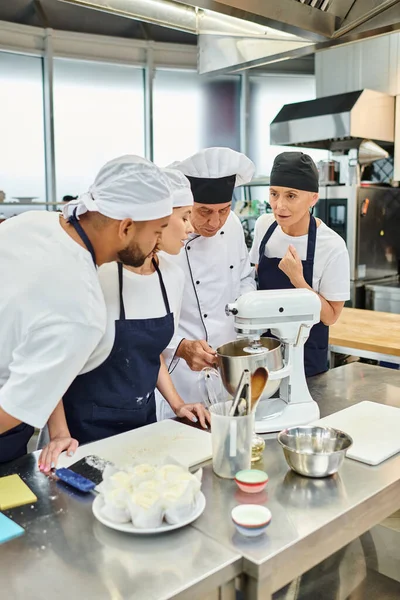 Chefs multirraciales bien parecido en delantales viendo a su jefe de cocina mostrando cómo trabajar con la batidora - foto de stock