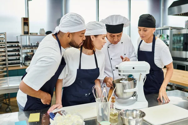 Multikulti-Köche in blauen Schürzen und Hauben beobachten ihren Chefkoch bei der Arbeit mit dem Mixer — Stockfoto
