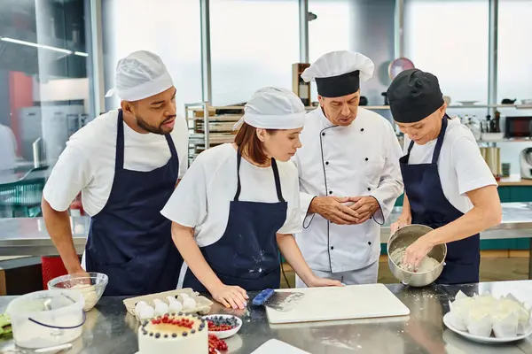 Young man and woman in aprons watching with their chief cook how their colleague working with dough — Stock Photo