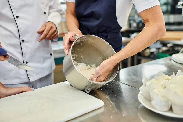 Cropped view of mature female chef in blue apron working with dough next to her chief cook — Stock Photo