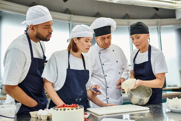 Beau chef cuisinier mature en chapeau blanc regarder comment ses divers chefs dans les tabliers de travail avec de la pâte — Photo de stock