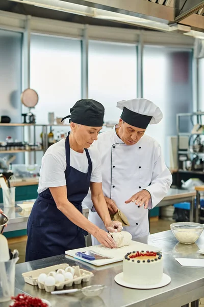 Chef maduro atraente em avental e toque trabalhando com massa ao lado de seu cozinheiro chefe, confeitaria — Fotografia de Stock