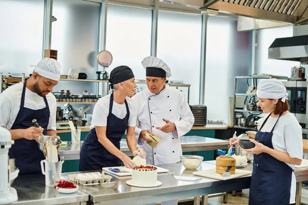 Mature chief cook in white hat talking to his chef next to her colleagues while on kitchen — Stock Photo
