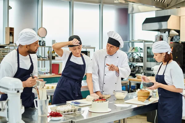 Cocinero jefe maduro en sombrero blanco explicando cómo hornear a sus chefs multiculturales que trabajan duro - foto de stock