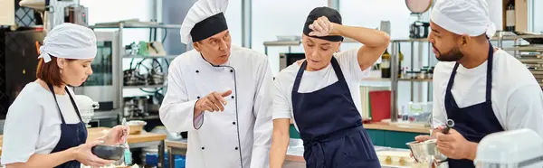 Cocinero jefe maduro en sombrero blanco que explica cómo hornear a sus chefs multirraciales que trabajan duro, bandera - foto de stock