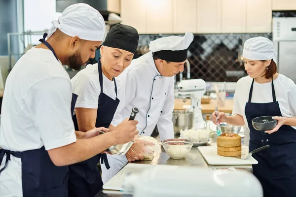 Chefs multiculturais em aventais azuis e toques trabalhando duro em sua pastelaria juntos, confeitaria — Fotografia de Stock