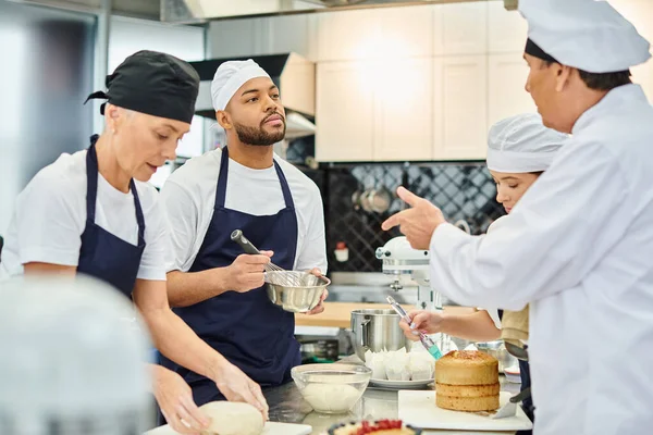 Mature chief cook talking to african american young chef while his colleagues working on pastry — Stock Photo