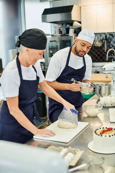 Focus on young african american chef in toque whisking next to his blurred jolly mature colleague — Stock Photo