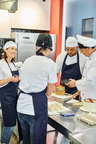 Jovem alegre em avental sorrindo para seus diversos colegas e chefe cozinheiro enquanto trabalhava em pastelaria — Fotografia de Stock
