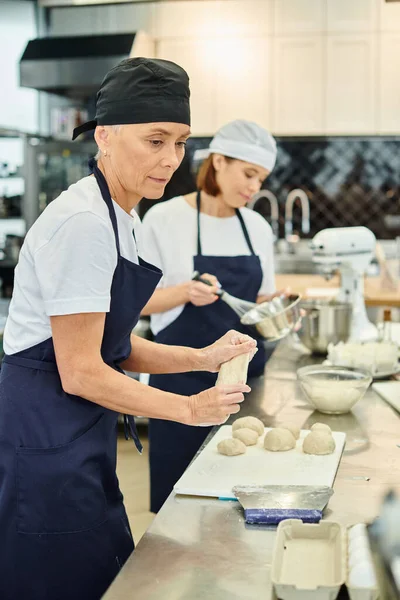 Focus on mature chef in toque and apron working with dough next to her young blurred colleague — Stock Photo