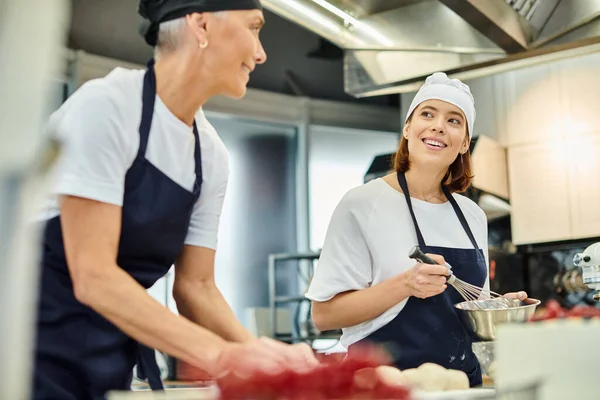 Foque em jovem chef alegre em avental sorrindo para seu colega maduro borrado enquanto trabalhava em pastelaria — Fotografia de Stock