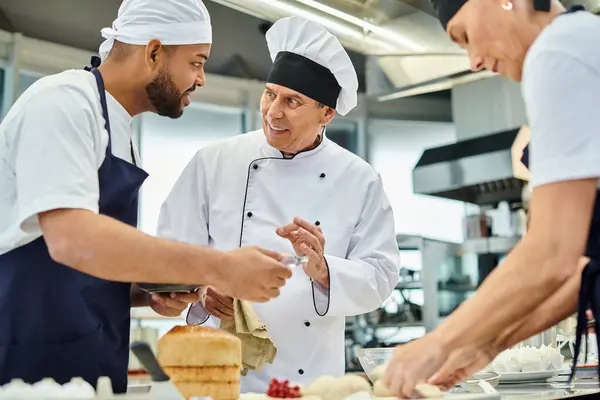 Alegre cocinero jefe maduro en sombrero blanco explicando la información alegremente a sus chefs multirraciales - foto de stock