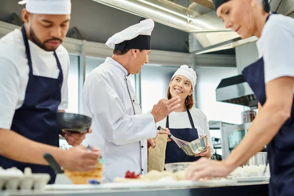 Alegre joven chef en toque sonriendo a su maduro jefe de cocina junto a sus diversos colegas - foto de stock