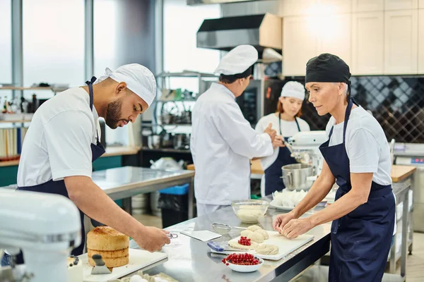 Focus on diverse chefs working with dough, their blurred colleague and chief cook baking on backdrop — Stock Photo
