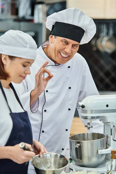 Centrarse en cocinero jefe alegre maduro en sombrero blanco mostrando gesto bien al lado de su chef femenina borrosa - foto de stock