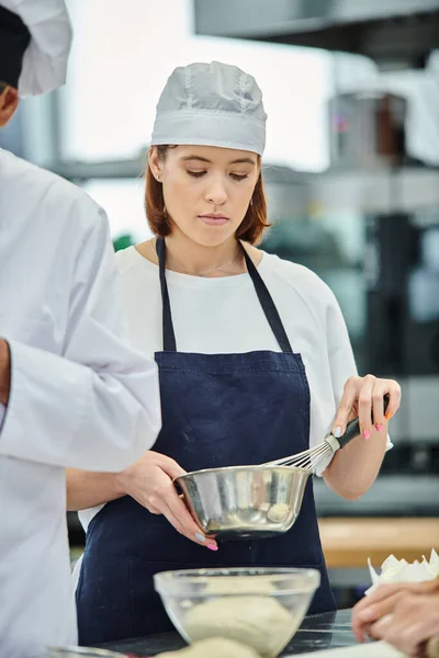 Attrayant jeune chef féminin en toque et tablier fouettant la pâte avant la cuisson, confiserie — Photo de stock