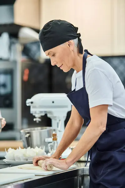 Mature gaie femme chef en toque bleue et tablier de travail avec de la pâte sur la cuisine, confiserie — Photo de stock