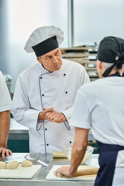 Bien parecido cocinero jefe maduro en sombrero blanco mirando atentamente al chef en la cocina, confitería - foto de stock