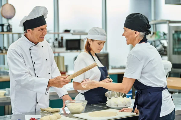 Beau chef cuisinier joyeux donnant rouleau à pâtisserie à son chef féminin à côté de leur jeune collègue — Photo de stock