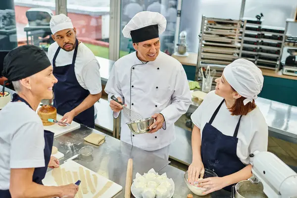Guapo jefe de cocina alegre en sombrero blanco rodeado de sus chefs multirraciales, confitería - foto de stock
